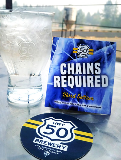 Icy glass of clear seltzer, "Chains Required" on table with view of trees behind.